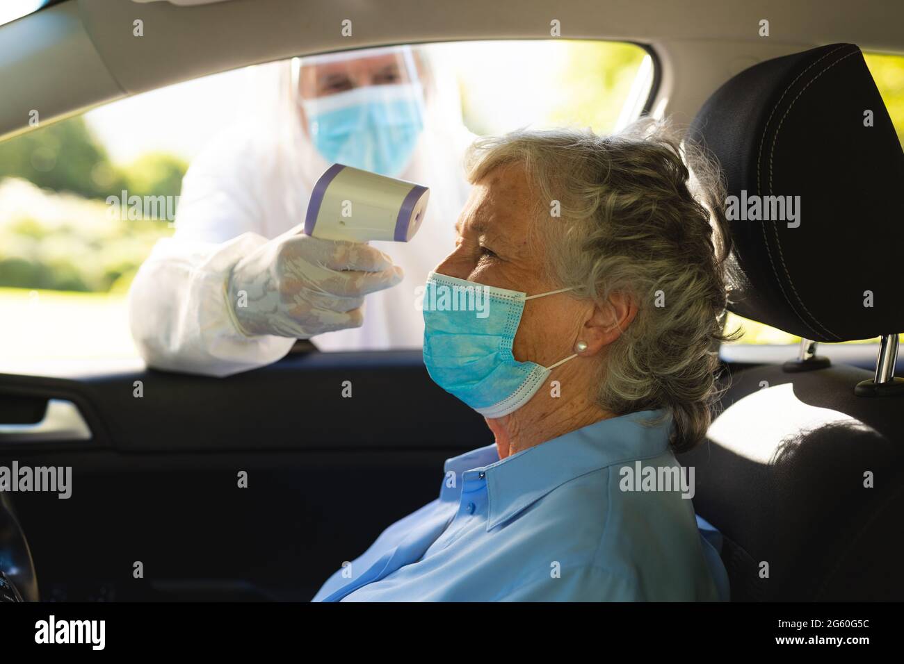 Medical worker wearing ppe suit taking temperature of senior caucasian woman sitting in car Stock Photo