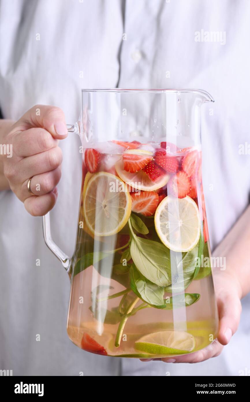 A jug with a refreshing organic soft drink with ripe fresh strawberries, lemon slices with lime and basil, in female hands close-up. Stock Photo