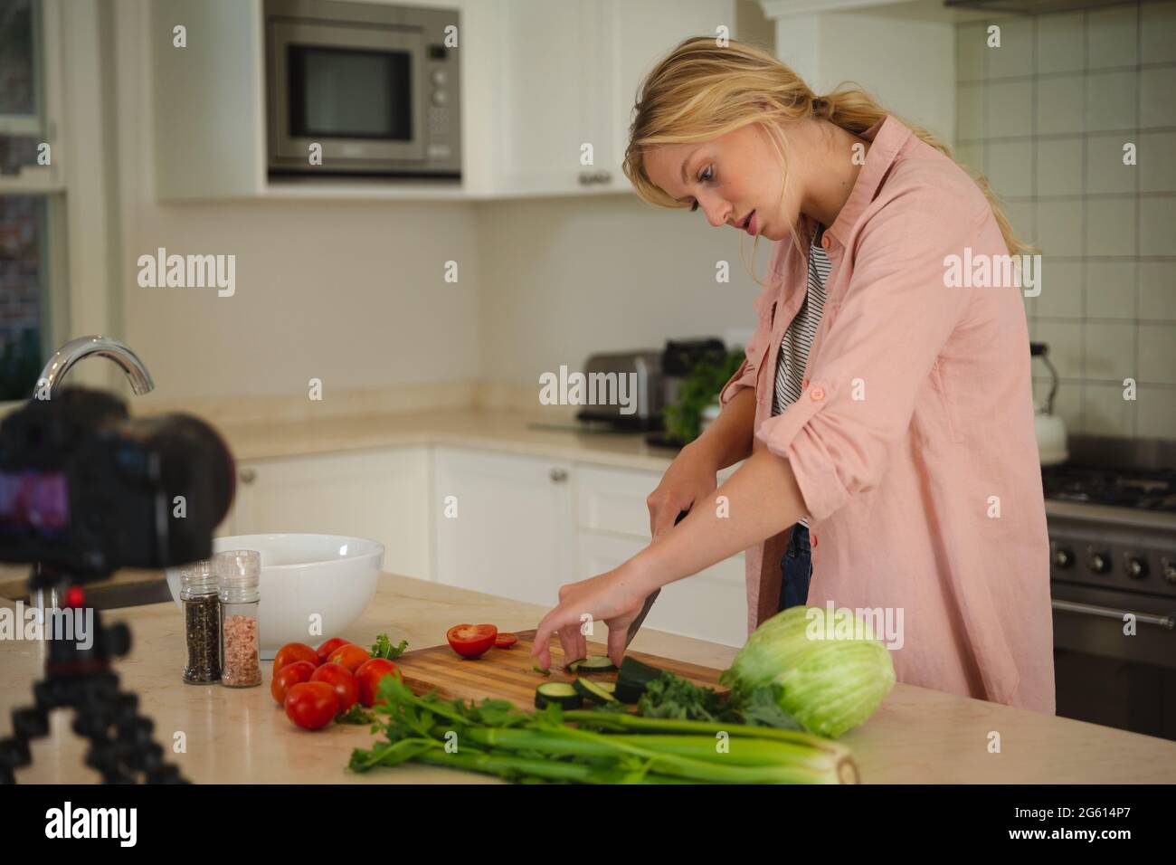 Caucasian woman in kitchen chopping vegetables and using camera, making cooking vlog Stock Photo
