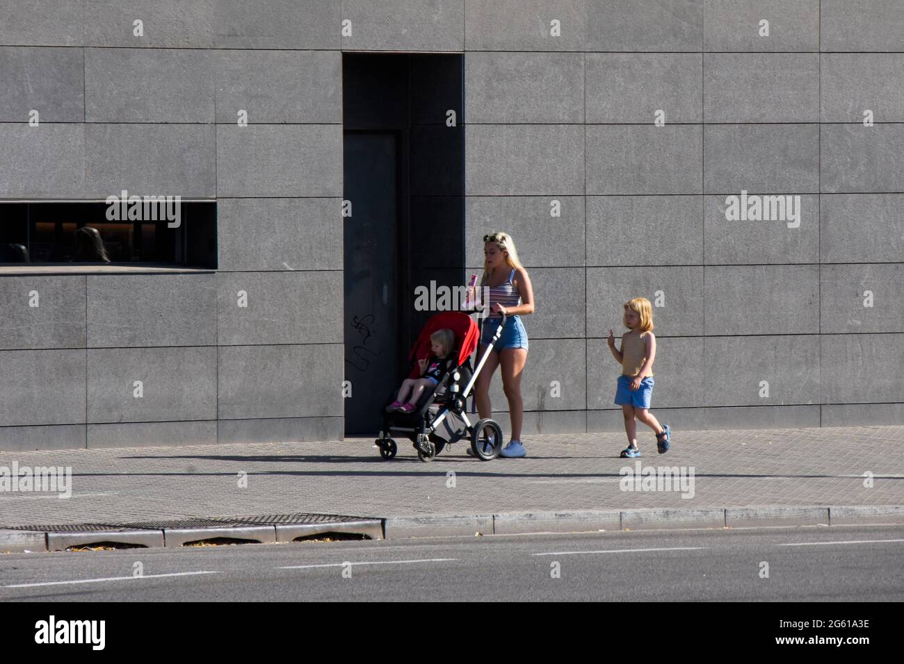 Family eating ice cream down the street. Mother with her two little ones. Blond woman with ice cream pushing the stroller with her daughter inside. Stock Photo