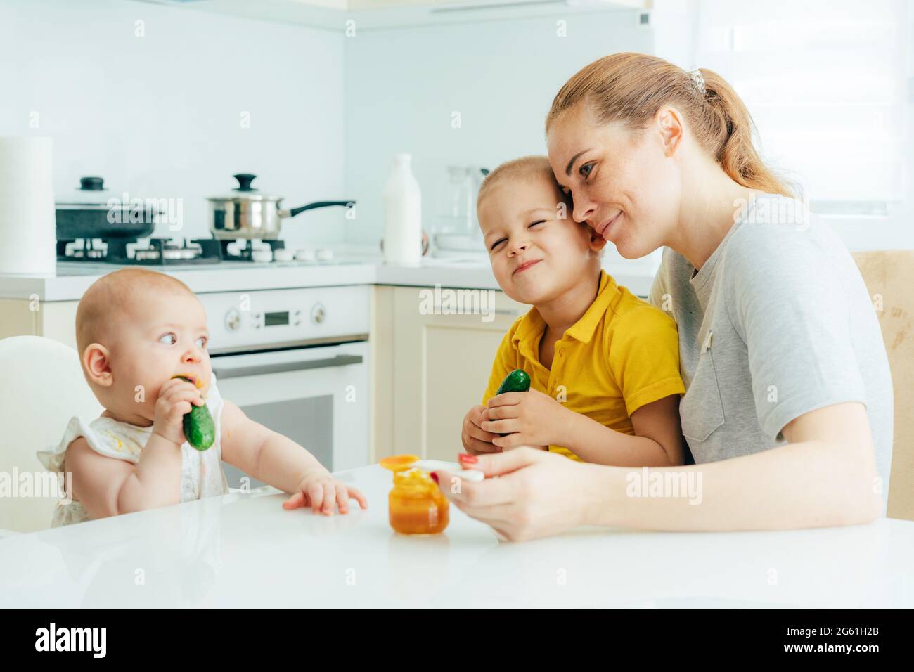 Candid family is at home in the kitchen, mom feeds the baby sitting in a chair, the eldest son sits on mom's lap and hugs her. Stock Photo