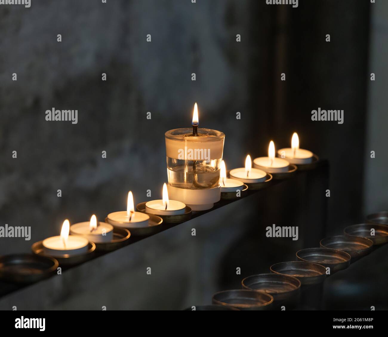 June 21st 2021: Votive candles. In a dark English church a large candle with oil and a wick stands in the middle of a line of eight lit tea candles. Stock Photo