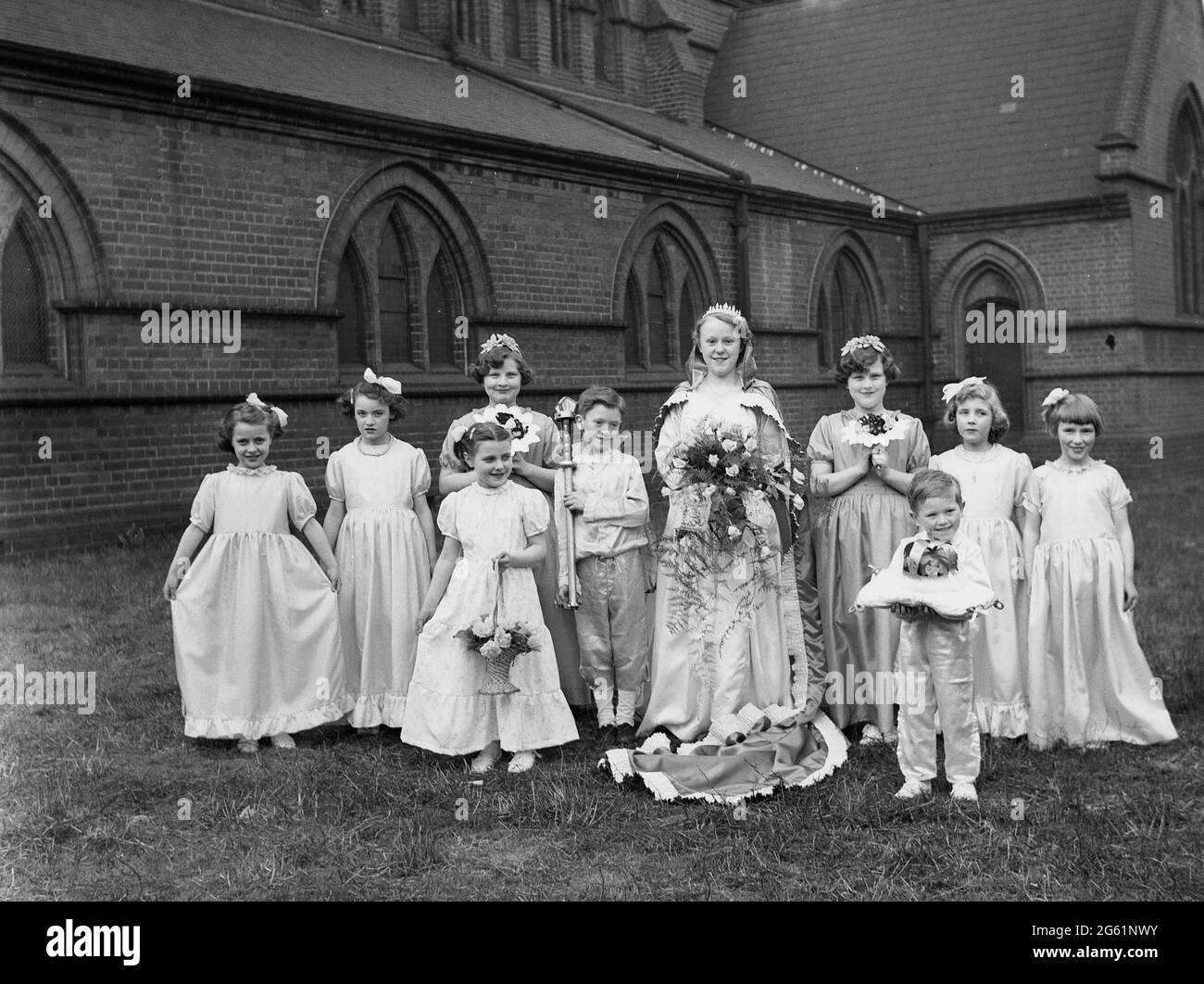 1956, historical, outside in the grounds of a church, a May Queen standing for a photo in her long gown and holding a bunch of flowers, with her entourage of helpers, young girls and two little boys, one of whom is holding a cushion with her crown on. A festival of ancient origins, held to celebrate the arrival of summer, May Day has been held in villages across Britain, with the crowning of the May Queen, a girl and a parade where she shows off her gown and crown to the local population. Stock Photo