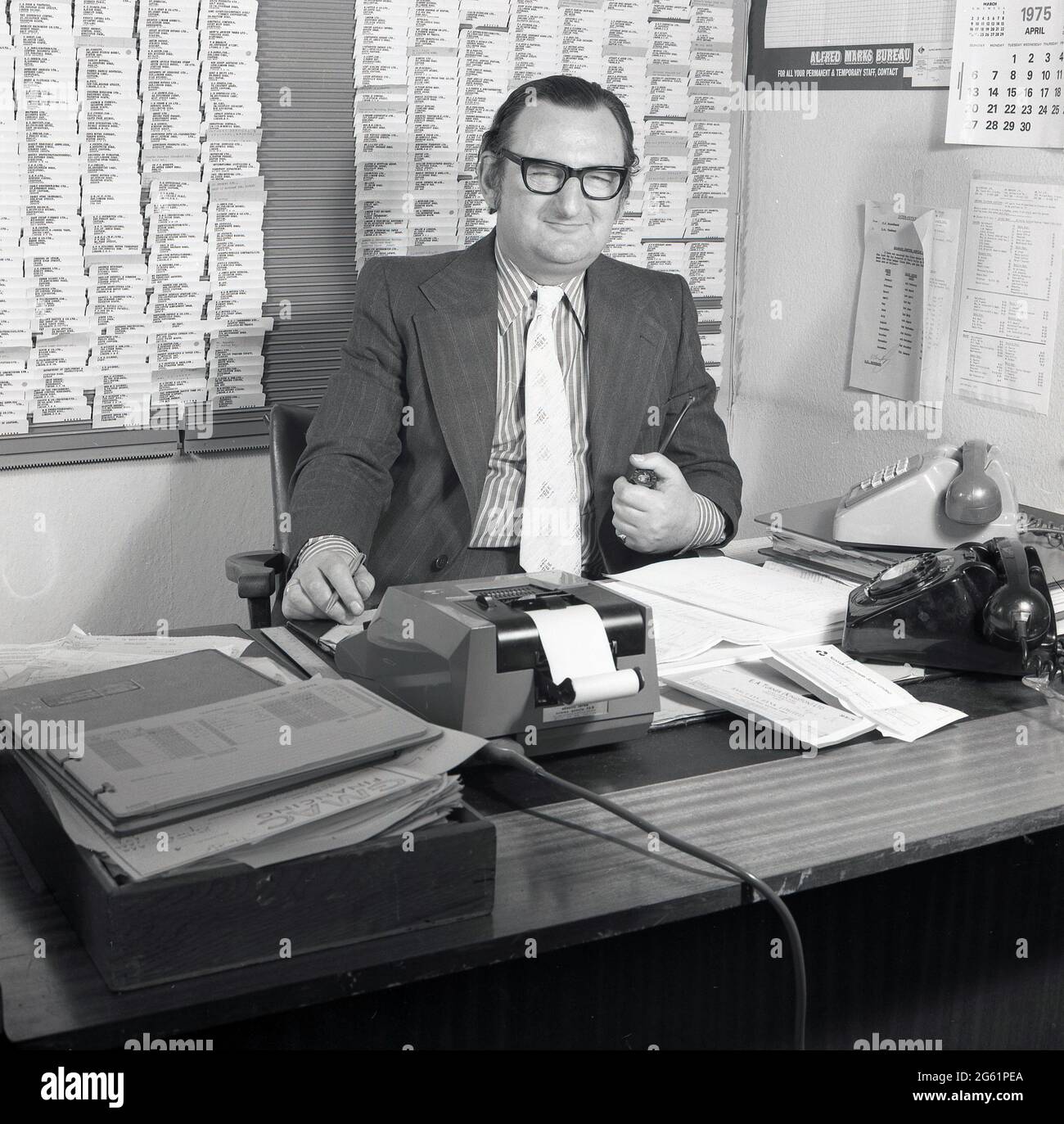 1970s, historical, inside an office, sitting at his desk, pipe in hand, a suited executive working in auto finance, with two telephones, paperwork and a mechanical adding machine around him, Croydon, England, UK. A walled customer/supplier card index is on the wall behind him. The financing of motor cars is big business. GMAC, an acronym for General Motors Acceptance Corporation was founded in 1919 and was part of the US car giant, General Motors, who owned the Vauxhall car brand in the UK, which they had acquired in 1925. Stock Photo