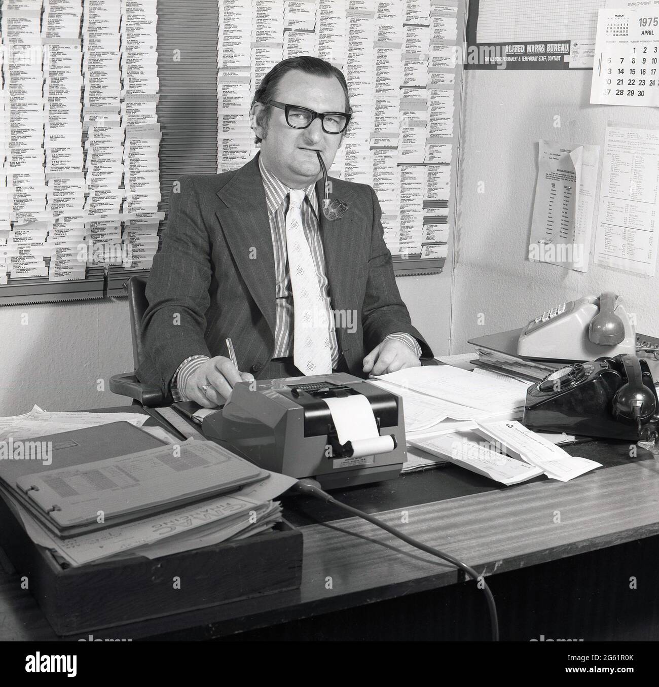 1970s, historical, inside an office, sitting at his desk, parker pen in hand and smoking a pipe, a suited executive working in auto finance, with two telephones, paperwork and a mechanical adding machine around him, Croydon, England, UK. A walled customer/supplier card index is on the wall behind him. The financing of motor cars is big business. GMAC, an acronym for General Motors Acceptance Corporation was founded in 1919 and was part of the US car giant, General Motors, who owned the Vauxhall car brand in the UK, which they had acquired in 1925. Stock Photo