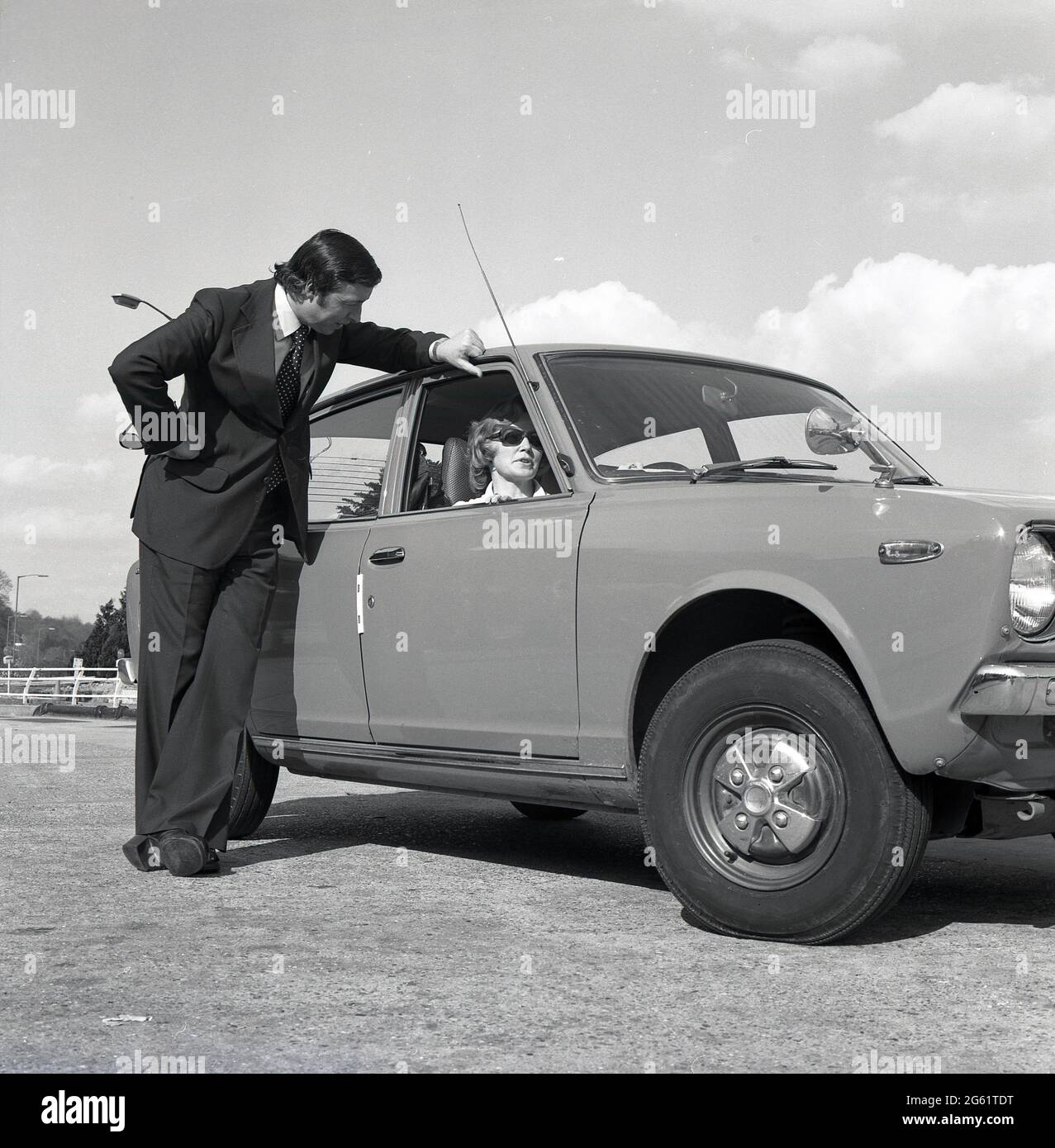 1975, on an off-road paved area, a man in a suit talking to an attractive woman in sunglasses sitting in her new Datsun Cherry 100A motor car, England, UK. Note the wide lapel of the man's suit jacket and flare bottoms of the trousers and the platform shoes, distinctive fashion styles of the '70s. Nissan began exporting Datsun-badged cars to the UK in 1968 and the Japanese brand was very successful, with well priced, reliable cars, at a time when the British motor industry was in a crisis, with  strikes by the workforce and a reputation for making cars of a poor quality and reliability. Stock Photo