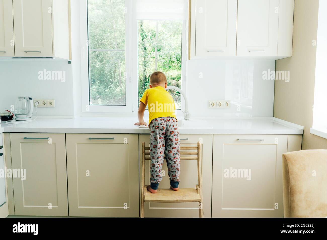 Little boy standing on a chair washes the dishes in the sink in the kitchen, back view. Stock Photo