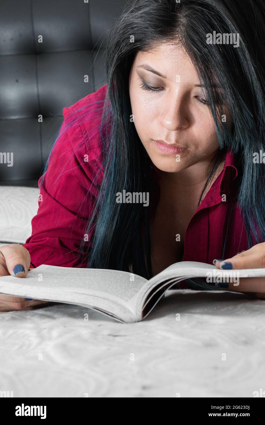blue haired woman lying on the bed, face down reading a book