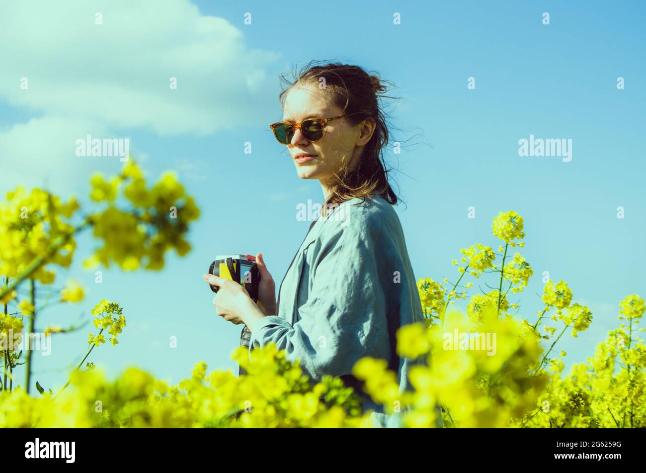 Bright vintage millennial portrait of a woman photographer with warrior hair bun in the middle of a yellow rapeseed field Stock Photo