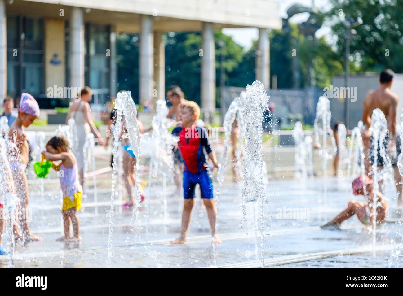Moscow, Russia, June 26, 2021. Selective focus on jets of water in a city fountain on a hot summer day. In the blurred background children and adults are batning and having fun. Stock Photo