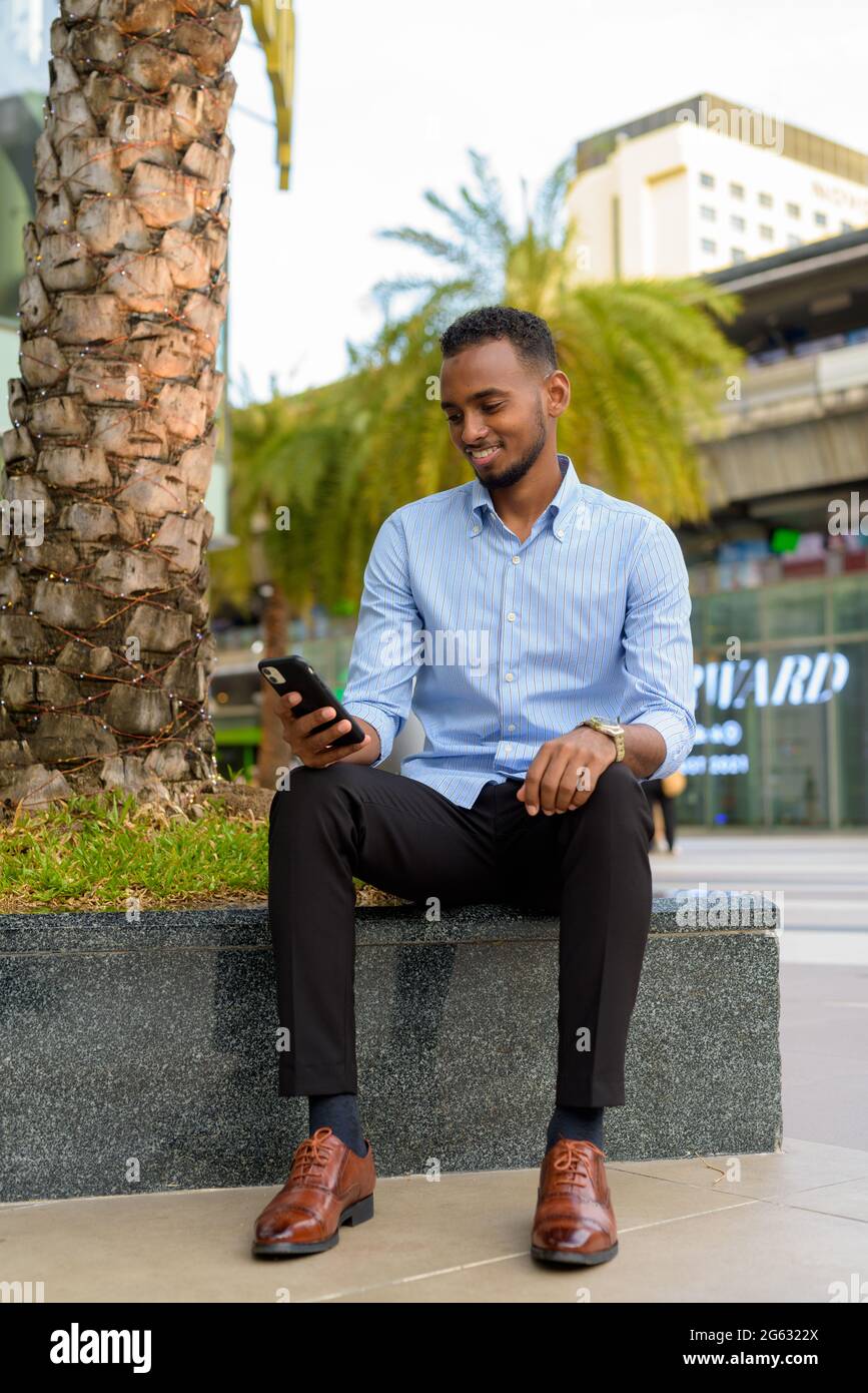 Portrait of handsome black African businessman outdoors in city during summer using mobile phone while smiling vertical shot Stock Photo