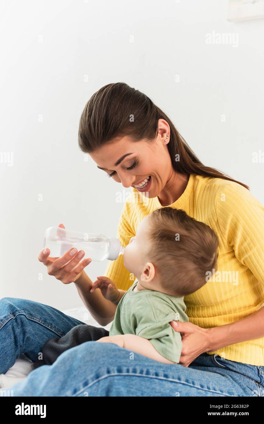 Smiling mother holding baby bottle while son drinking in bedroom Stock