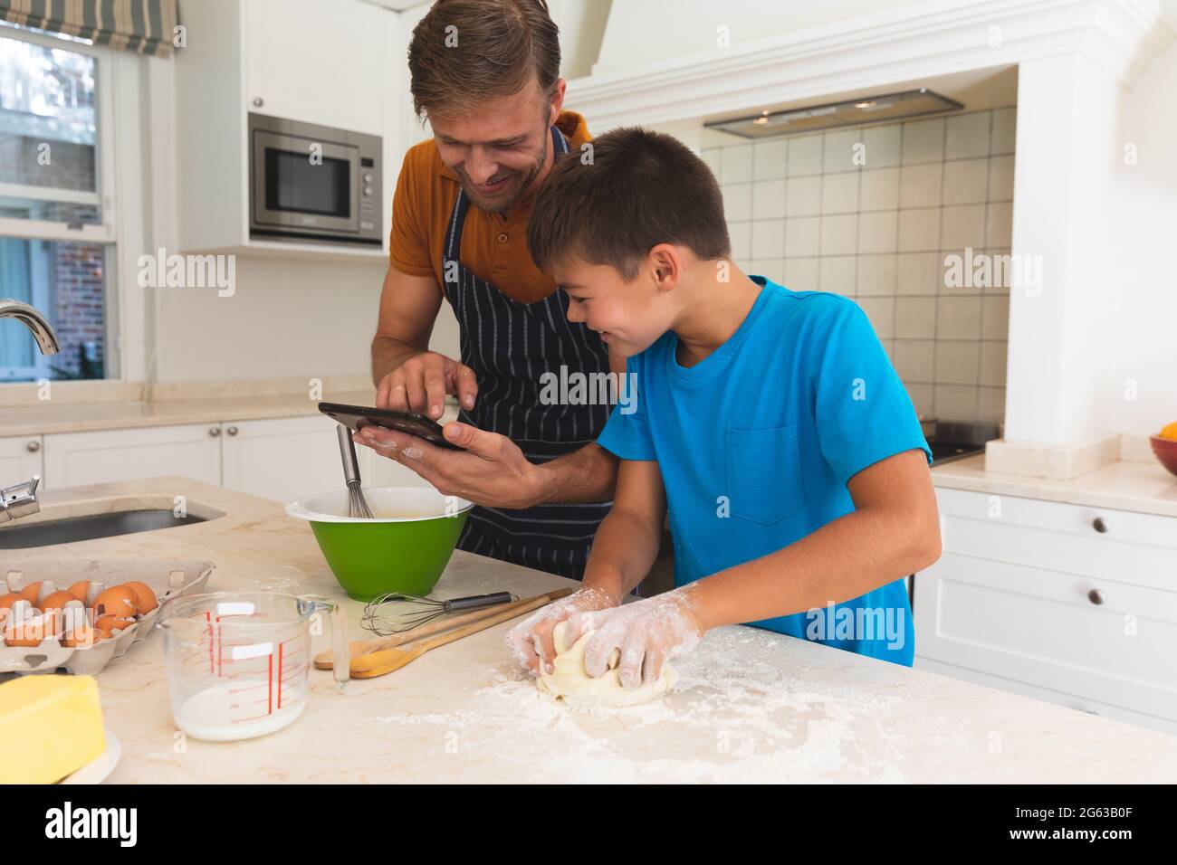 Caucasian father using tablet and son baking and smiling in kitchen Stock Photo