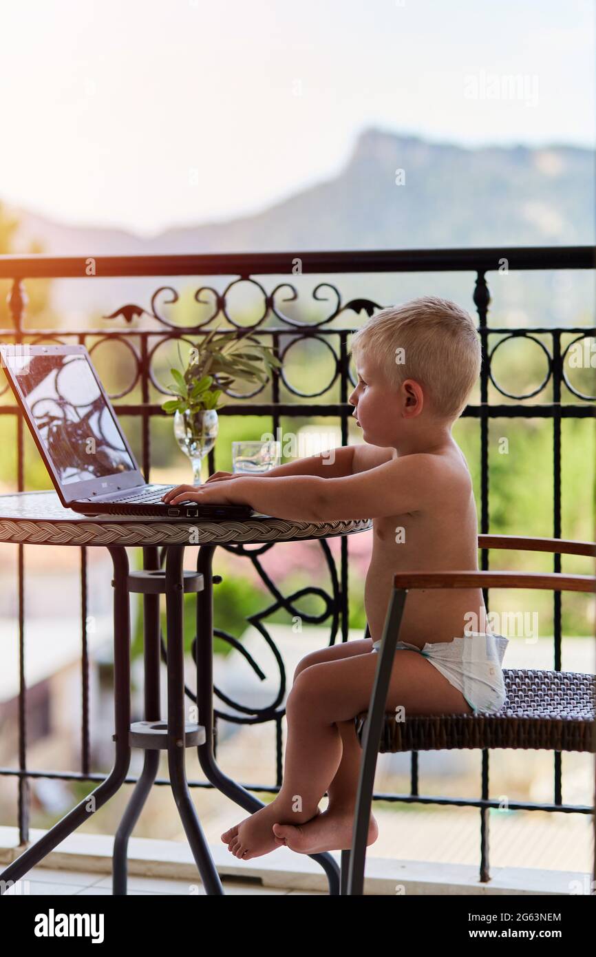 Caucasian toddler boy in diaper plays on laptop on terrace on sunny day. New generation of children. Playing games, watching cartoons in front of moun Stock Photo