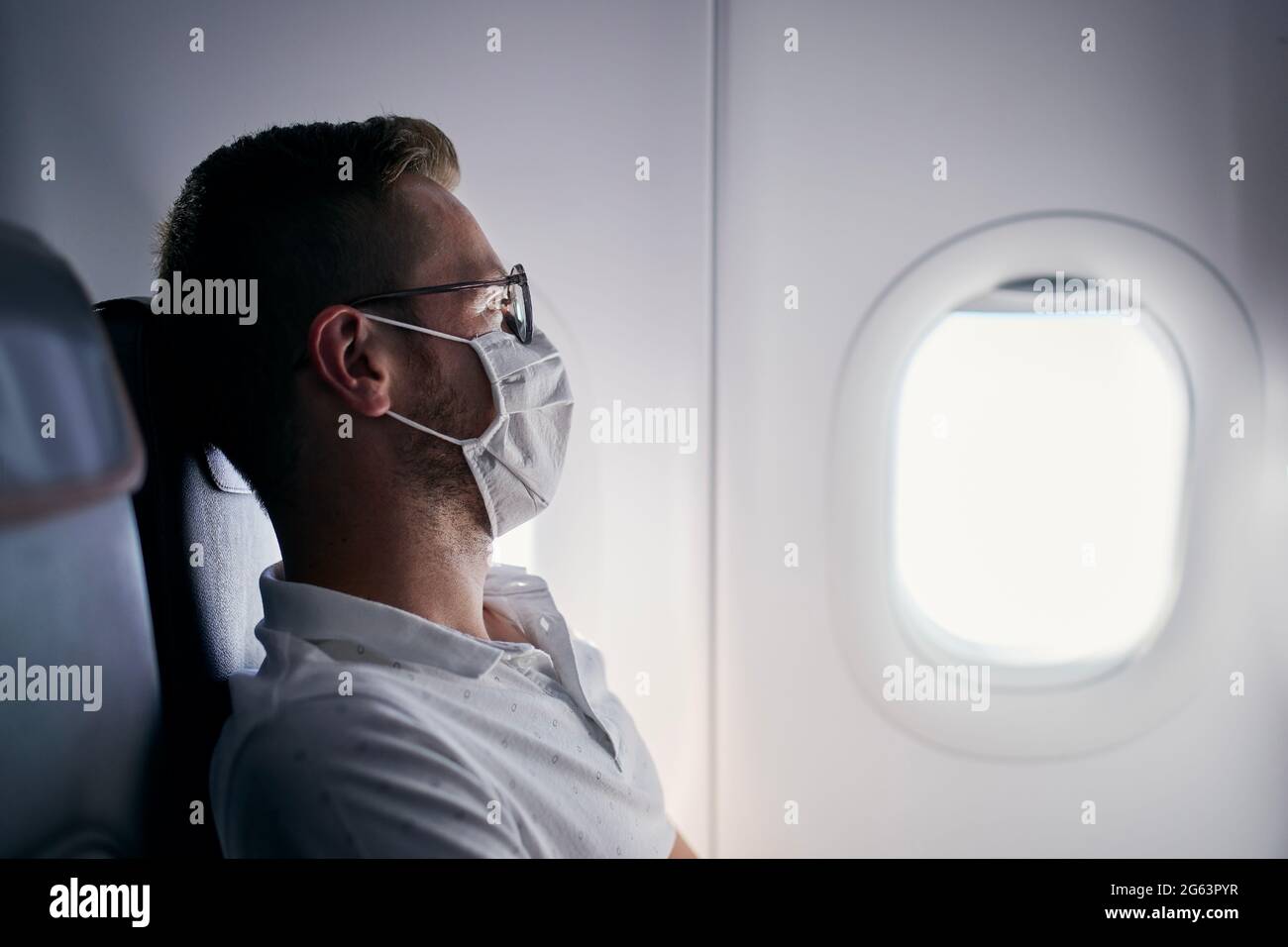 Passenger with protective face mask. Young man man looking out window of airplane. Themes traveling in new normal and personal protection during pande Stock Photo