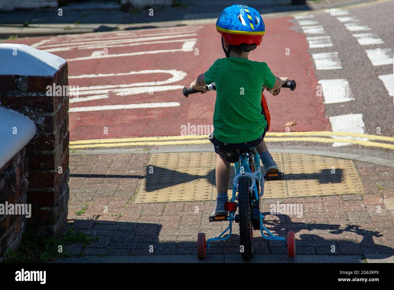 A small child riding a bike with stabilizers or stabailisers wearing a helmet, about to cross a road Stock Photo