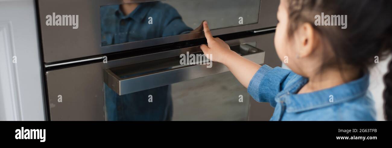 Asian kid on blurred foreground touching display of oven at home, banner Stock Photo