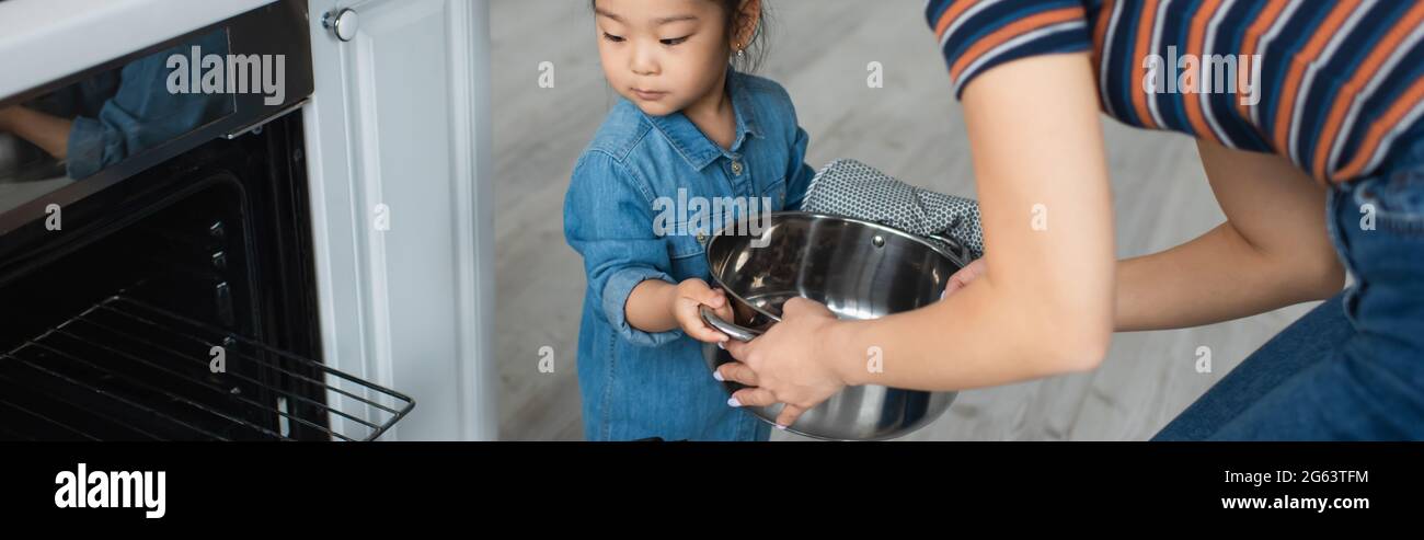 Mother holding pan near asian daughter and oven at home, banner Stock Photo