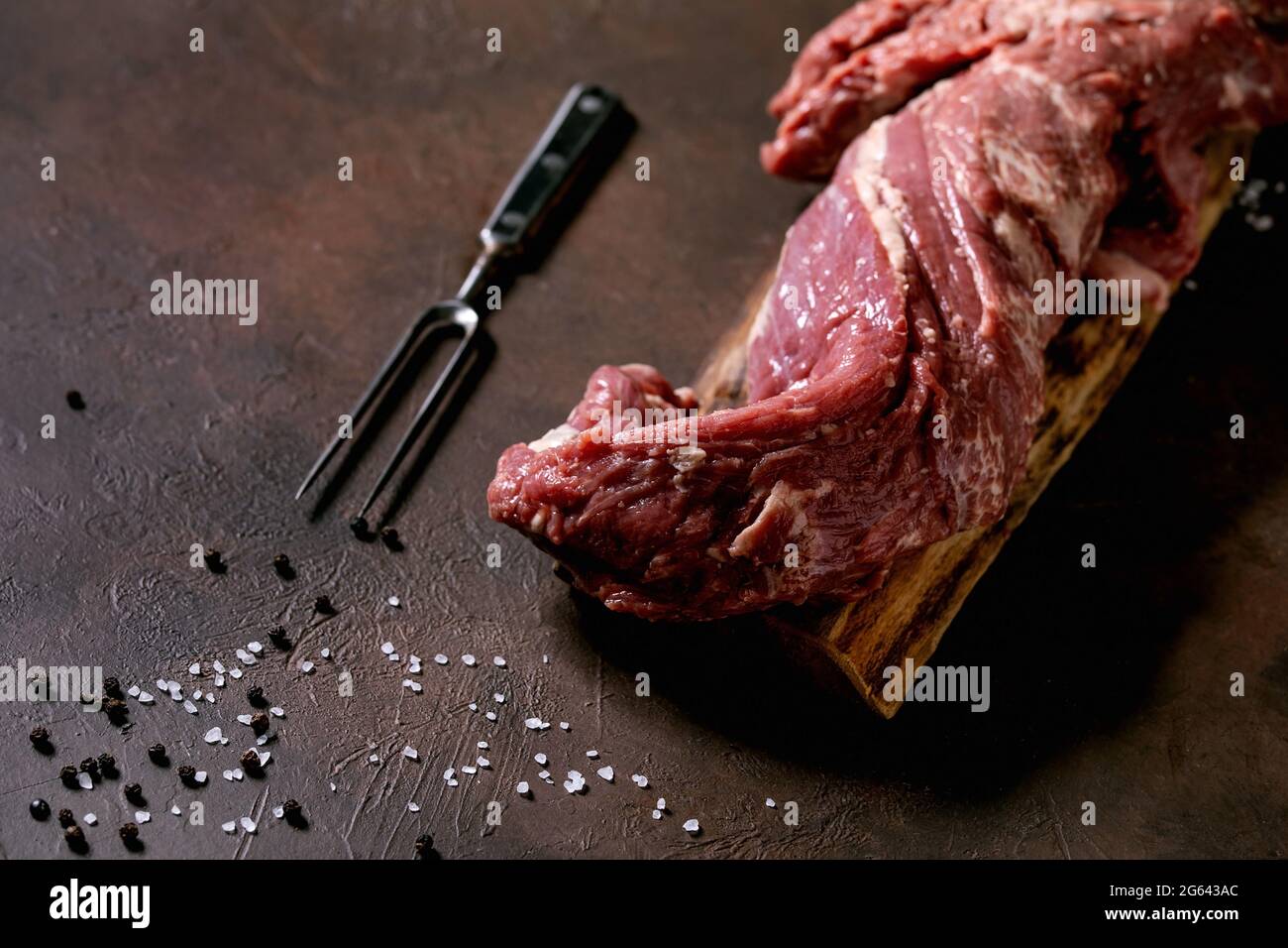 Fresh whole raw beef tenderloin meat on wooden board with metal meat fork, salt and pepper over dark brown texture background. Food cooking background Stock Photo