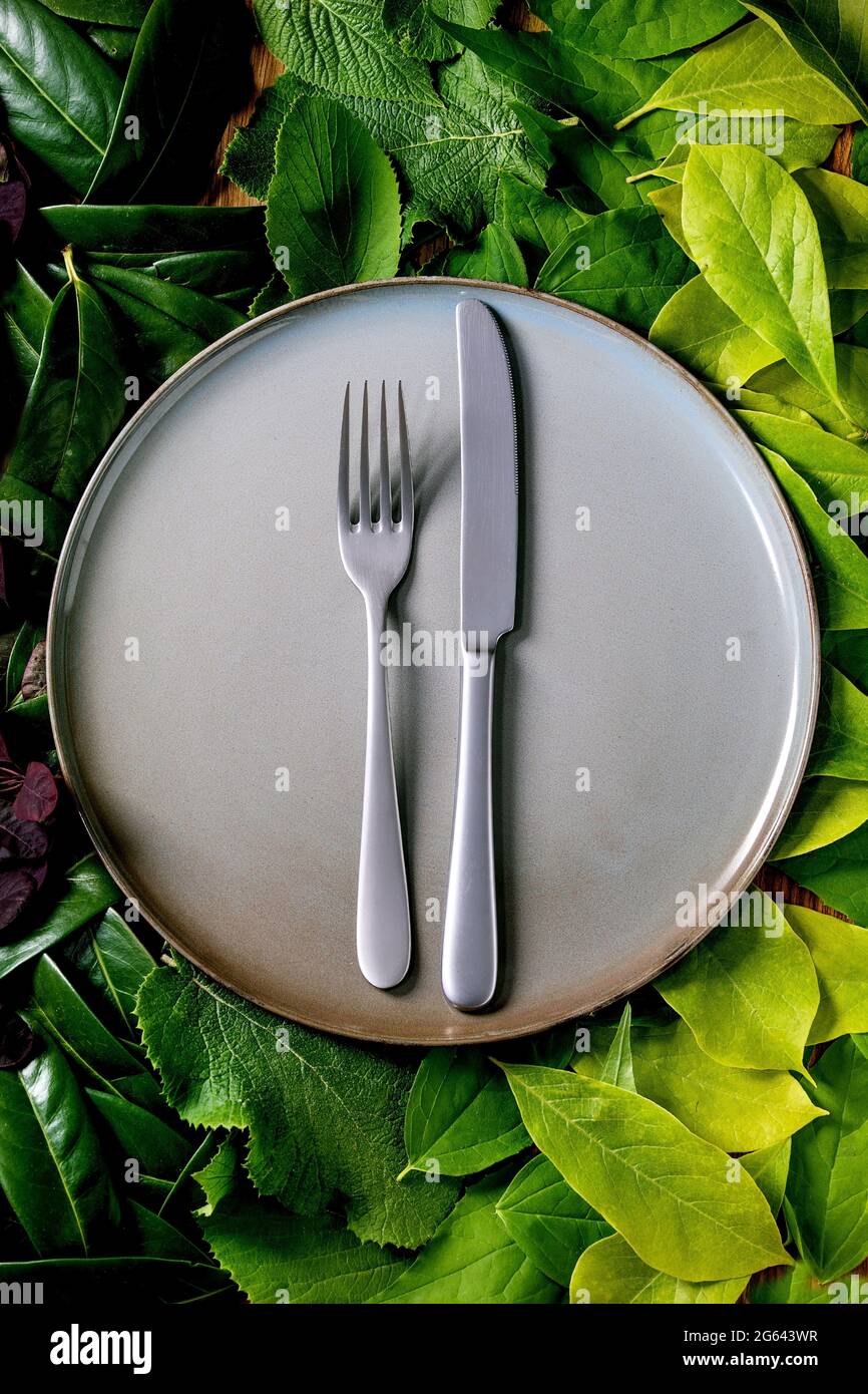 Empty ceramic plate with knife and fork on background made of green leaves, green gradient. Empty place for product. Summer menu eco food concept. Nat Stock Photo