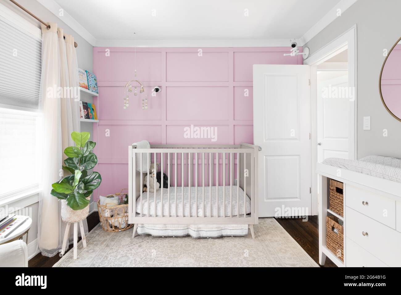 A cozy nursery with a crib in front of a light pink board and batten accent wall. A mobile hangs above the crib and cameras are mounted on the wall. Stock Photo