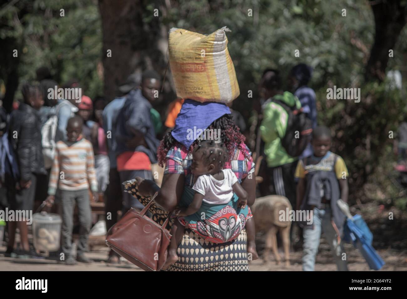 Mother carrying sleeping baby on a sarong cloth tied on her back and carrying a bag on her head Stock Photo