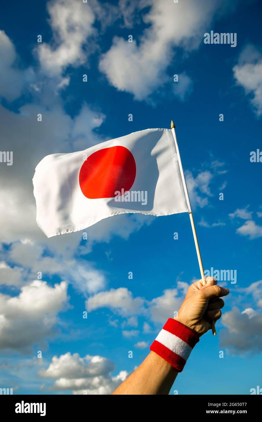 Hand with Japan red and white wristband holding a Japanese flag waving in bright sunny blue sky Stock Photo
