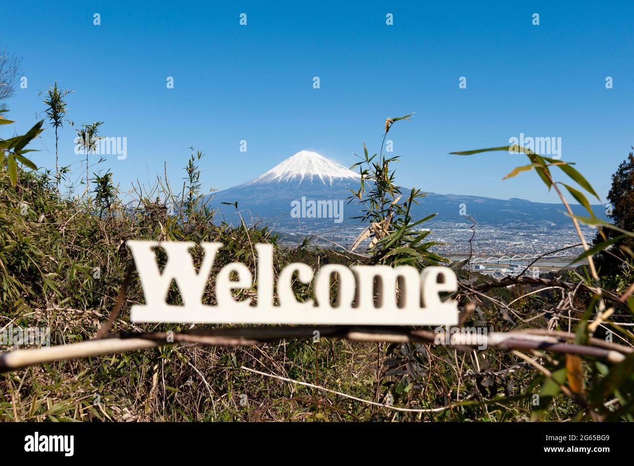 'Welcome' word written in wood. Background with Fuji City aerial view and the famous Mount Fuji with snowy peak and beautiful blue sky. Japan. Stock Photo