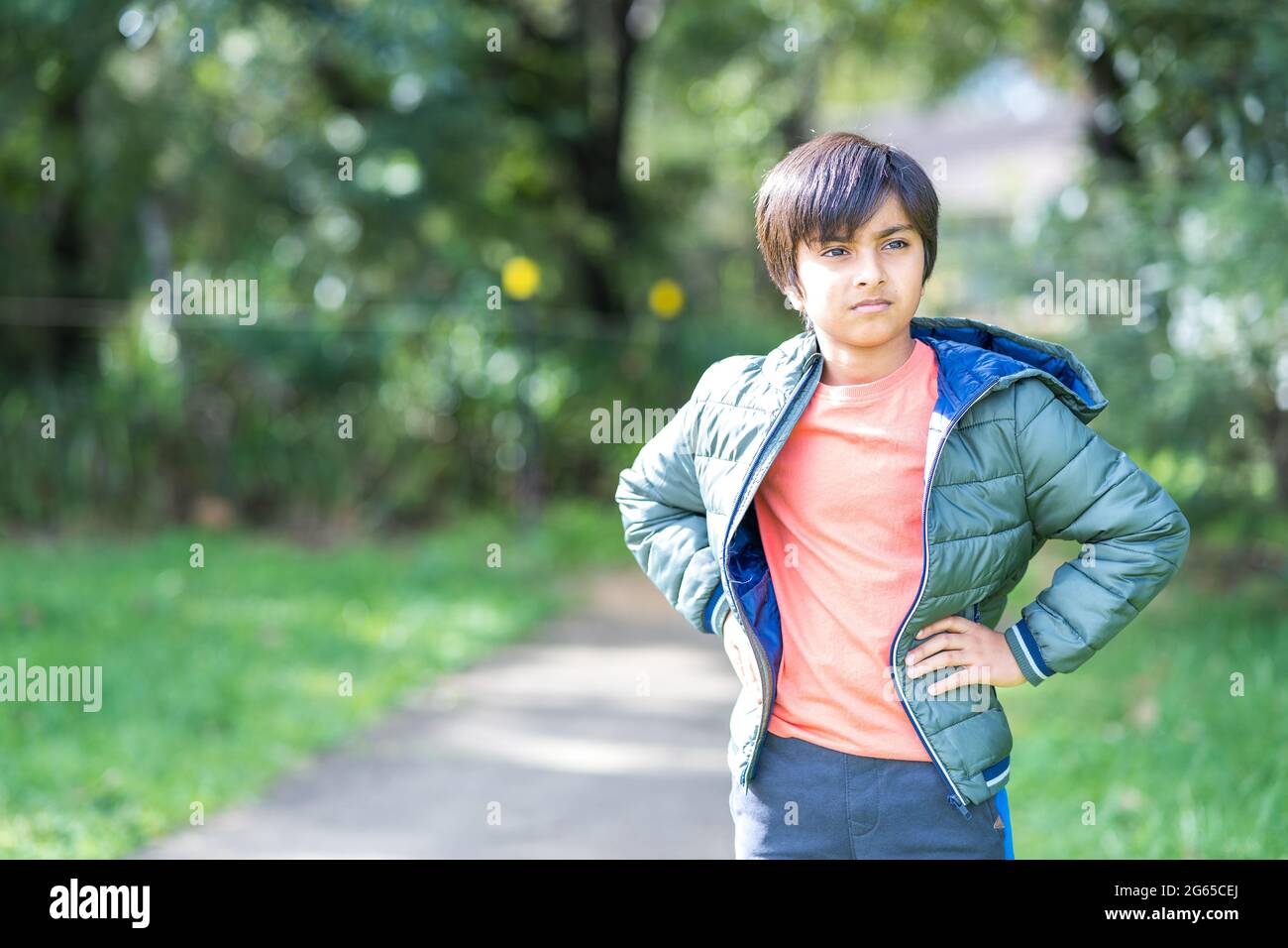 Portrait of tired young boy outdoors in natural light. South Asian child wearing puffer jacket with hands on waist looking in the distance. Stock Photo