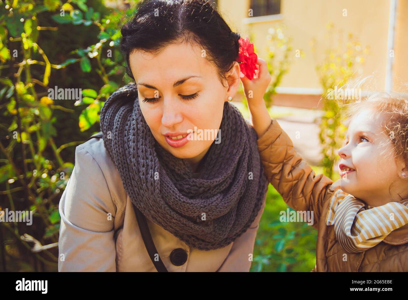 A little happy girl in a mustard-colored jacket puts red flower in mothers hair Stock Photo