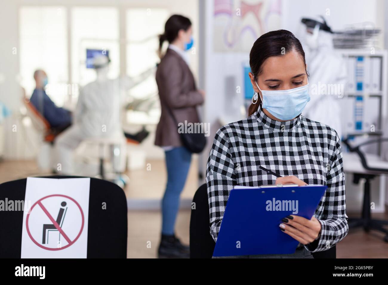 Stomatology patient in denstiry wainting area filling form before consultation with dentist dressed in ppe suit as safety precation agasint infection with coronavirus during global outbreak. Stock Photo