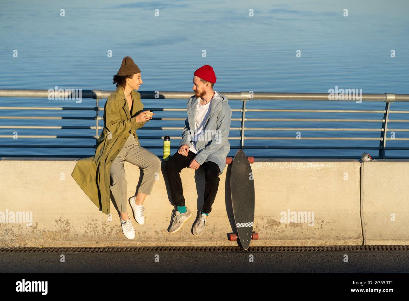 Young couple of hipsters enjoy time together sit on bridge at river drink hot tea from thermos talk Stock Photo