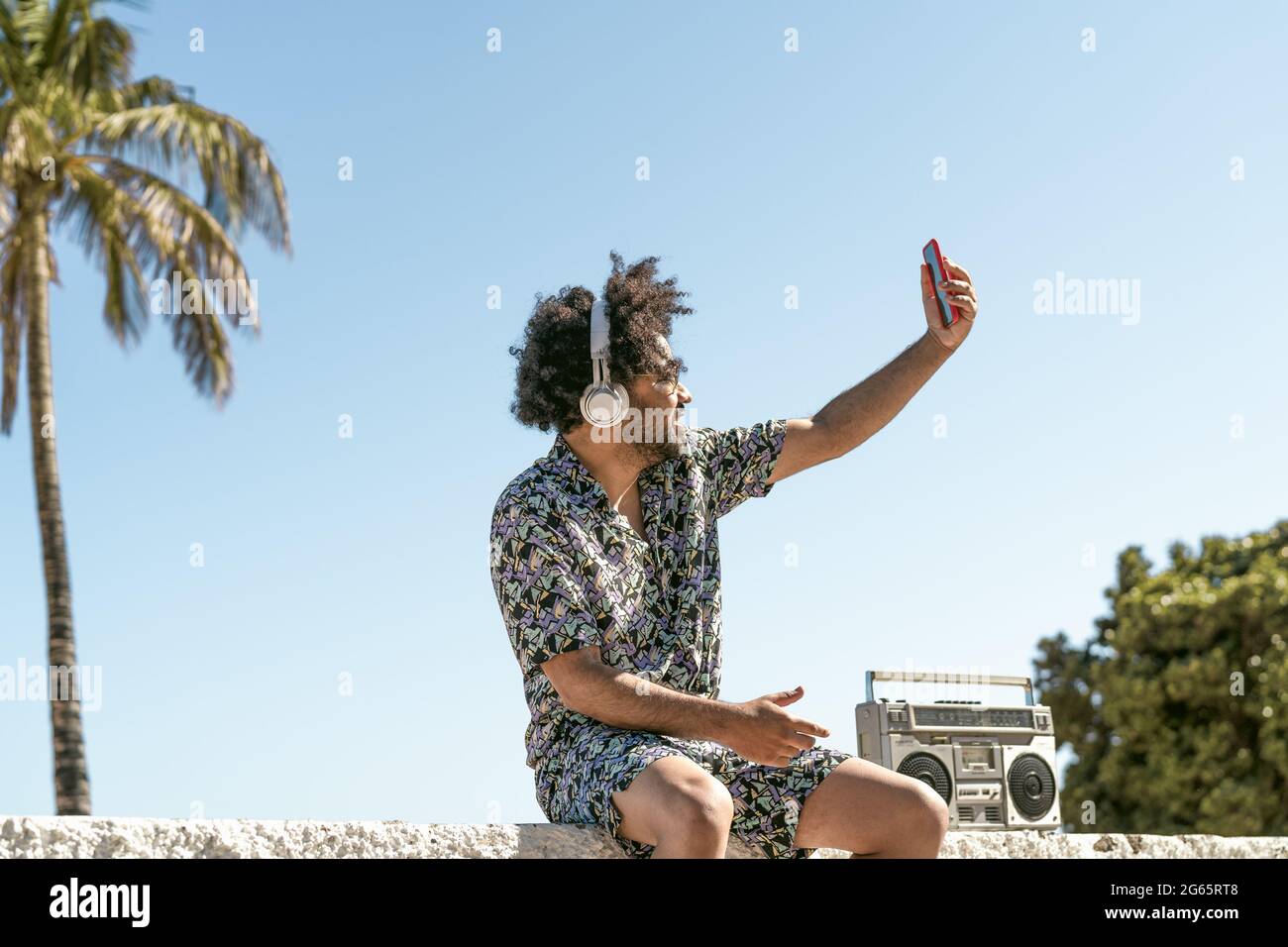 Young man having fun taking selfie with mobile smartphone while listening music with headphones and vintage boombox during summer vacations Stock Photo