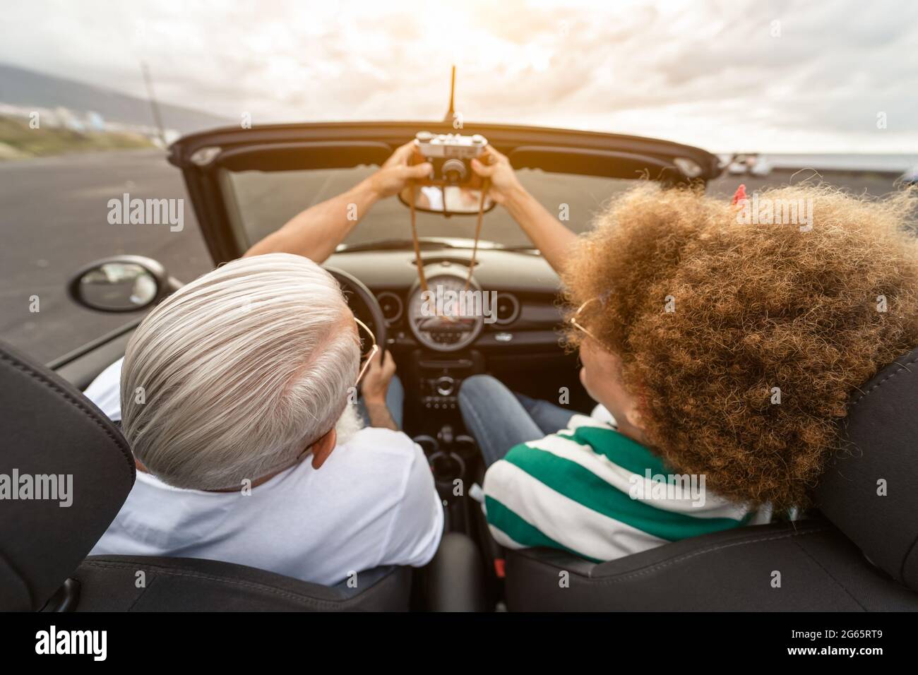 Happy senior couple having fun taking photo with old vintage camera while driving on new convertible car during road trip tour vacation Stock Photo