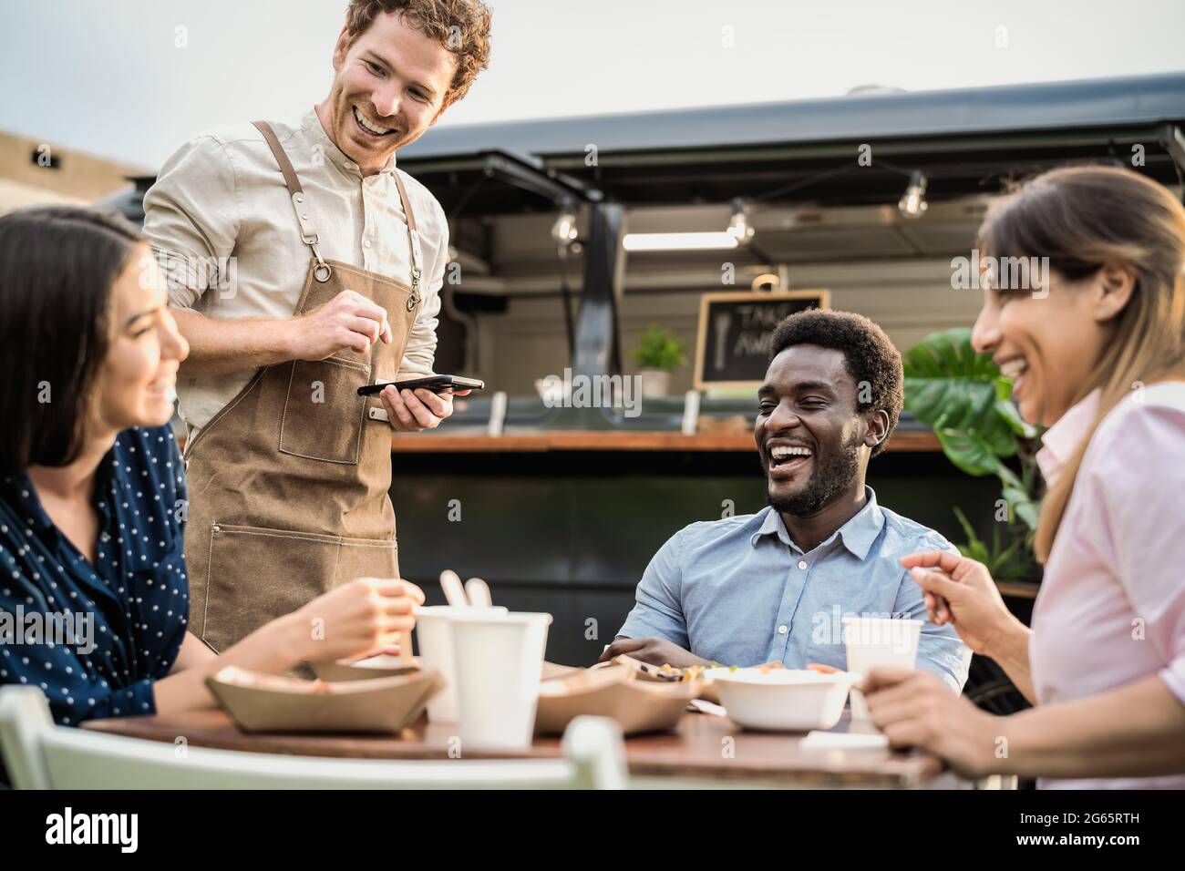 Food truck owner taking customers orders with mobile device - Happy multiracial people having a meal outdoor Stock Photo
