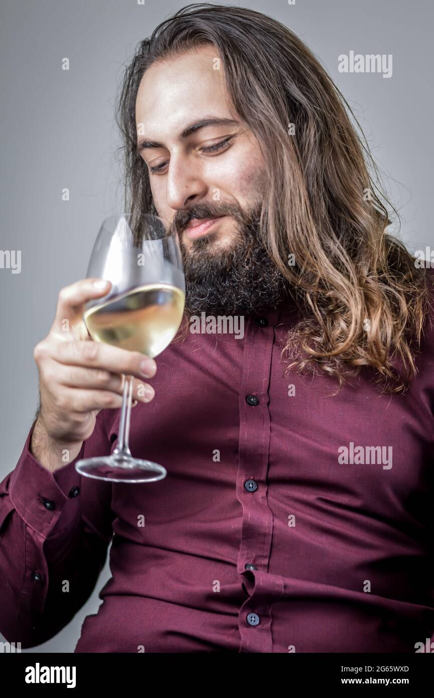 Portrait of a young Middle Eastern businessman with a beard and long hair sniffing a glass of white wine to smell its bouquet Stock Photo