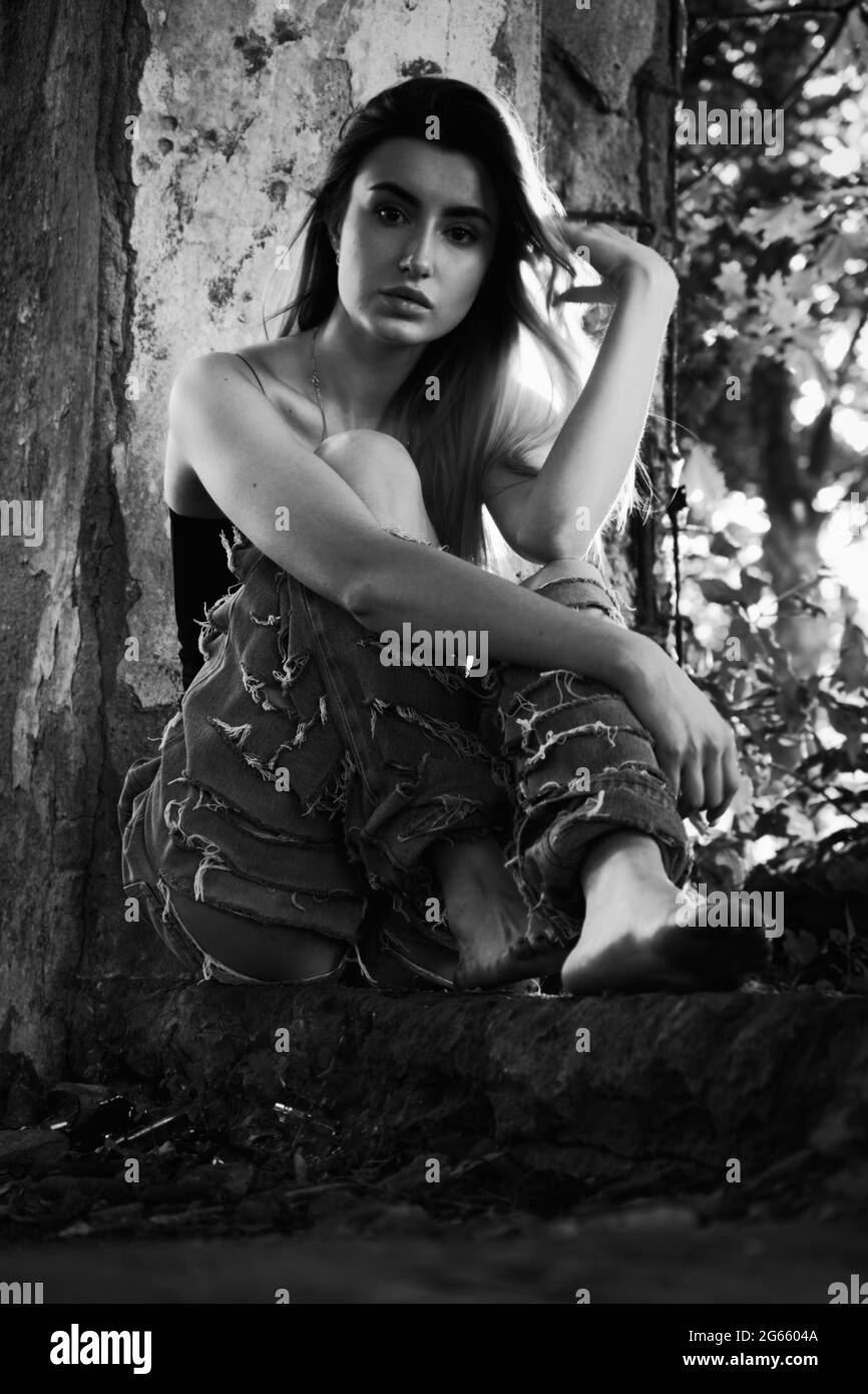 tramp girl in torn jeans sits in old dirty ruins, monochrome Stock Photo