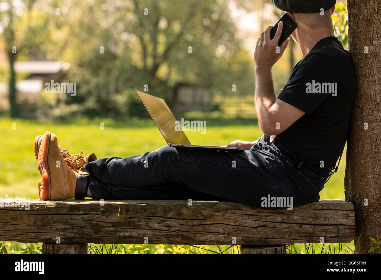 Farmer man wearing a cap and casual clothes sitting on a wooden bench calling with his mobile phone, working on his laptop and doing business while lo Stock Photo