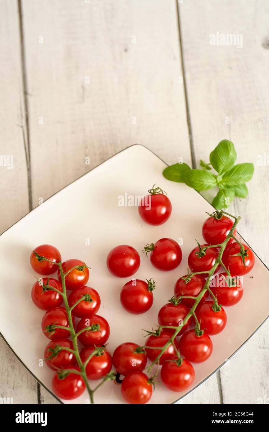 Red Cherry Tomatoes And Basil On A White Plate And White Wooden Table. Fehraltorf, Switzerland Stock Photo