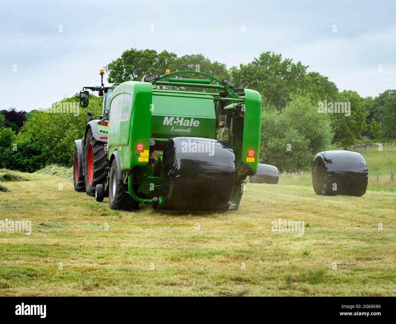 Hay or silage making (farmer & farm tractor at work in rural field, collecting grass, wrapped round bale dropping from baler) - Yorkshire England, UK. Stock Photo