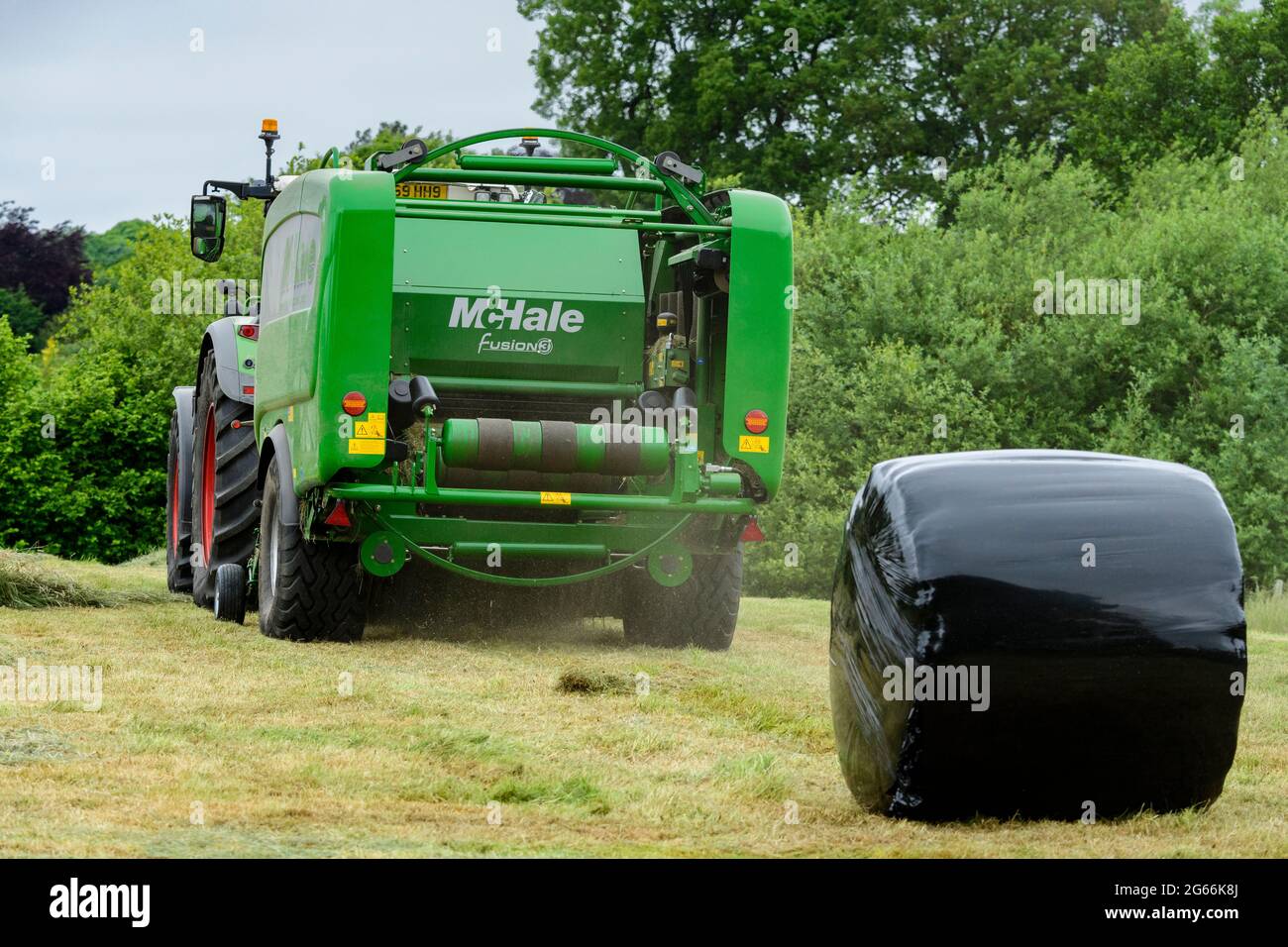 Hay or silage making (farmer in farm tractor at work in rural field, collecting dry grass, green baler & wrapped round bale) - Yorkshire, England UK. Stock Photo