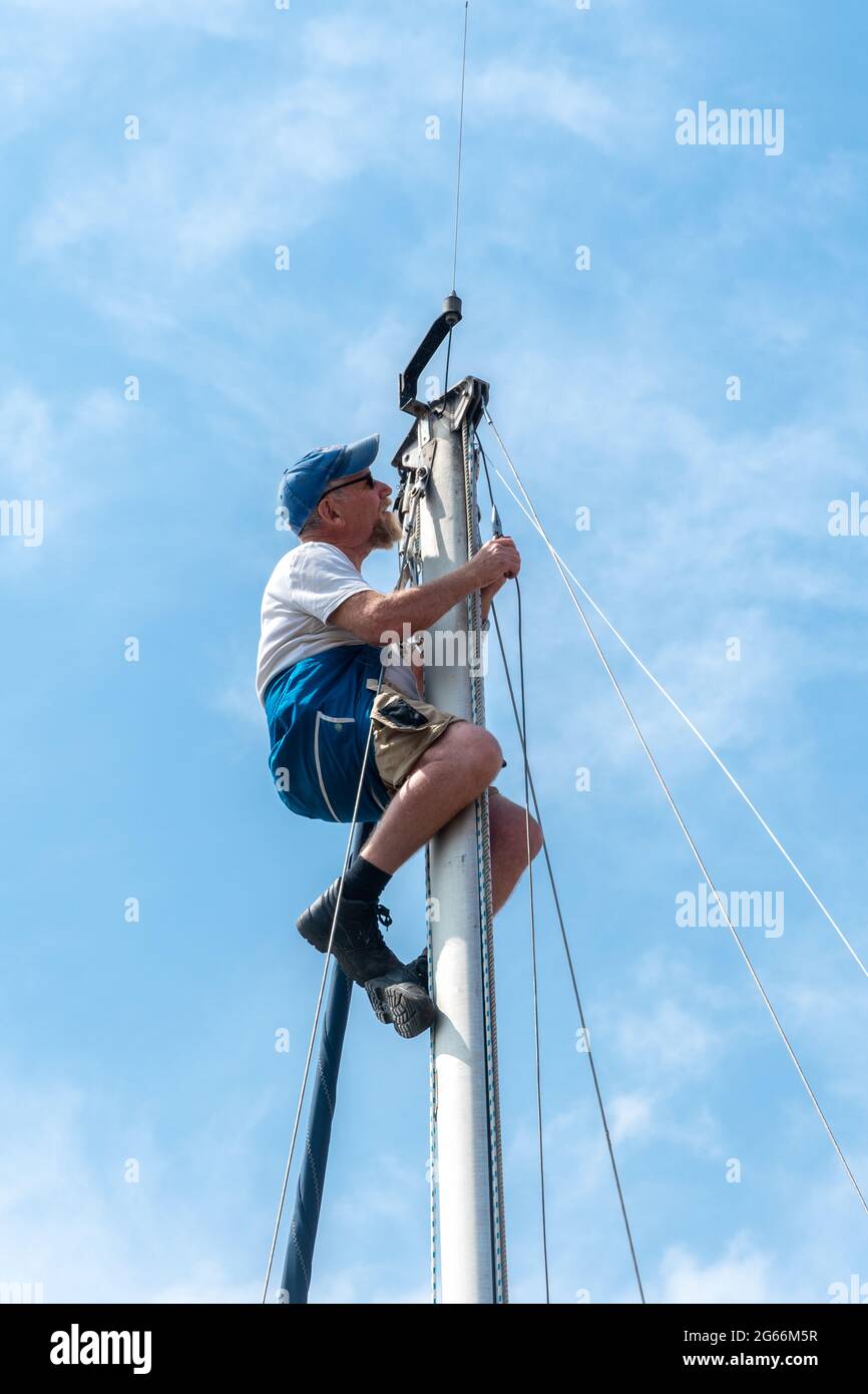 Man in a safety harness at the top of the mast of a yacht or sailboat carrying out maintenance work on the rigging, UK Stock Photo