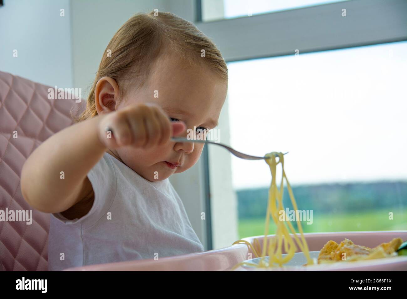 Little beautiful one-year-old girl in a white T-shirt eats pasta with a fork, sitting in a pink baby chair. Stock Photo