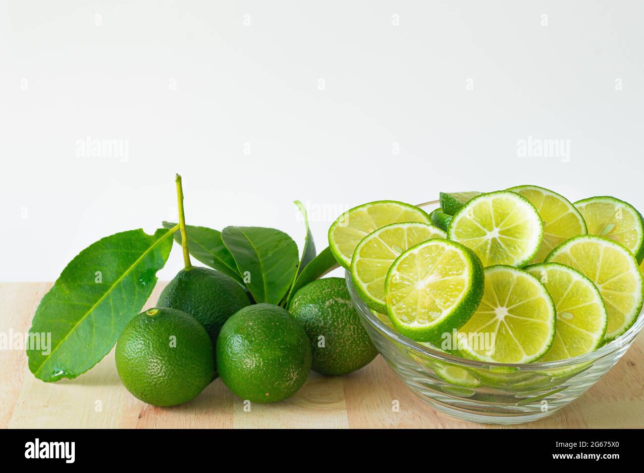 Green limes with leaves and sliced lime in glass bowl on wood, white background. Copy space. Stock Photo