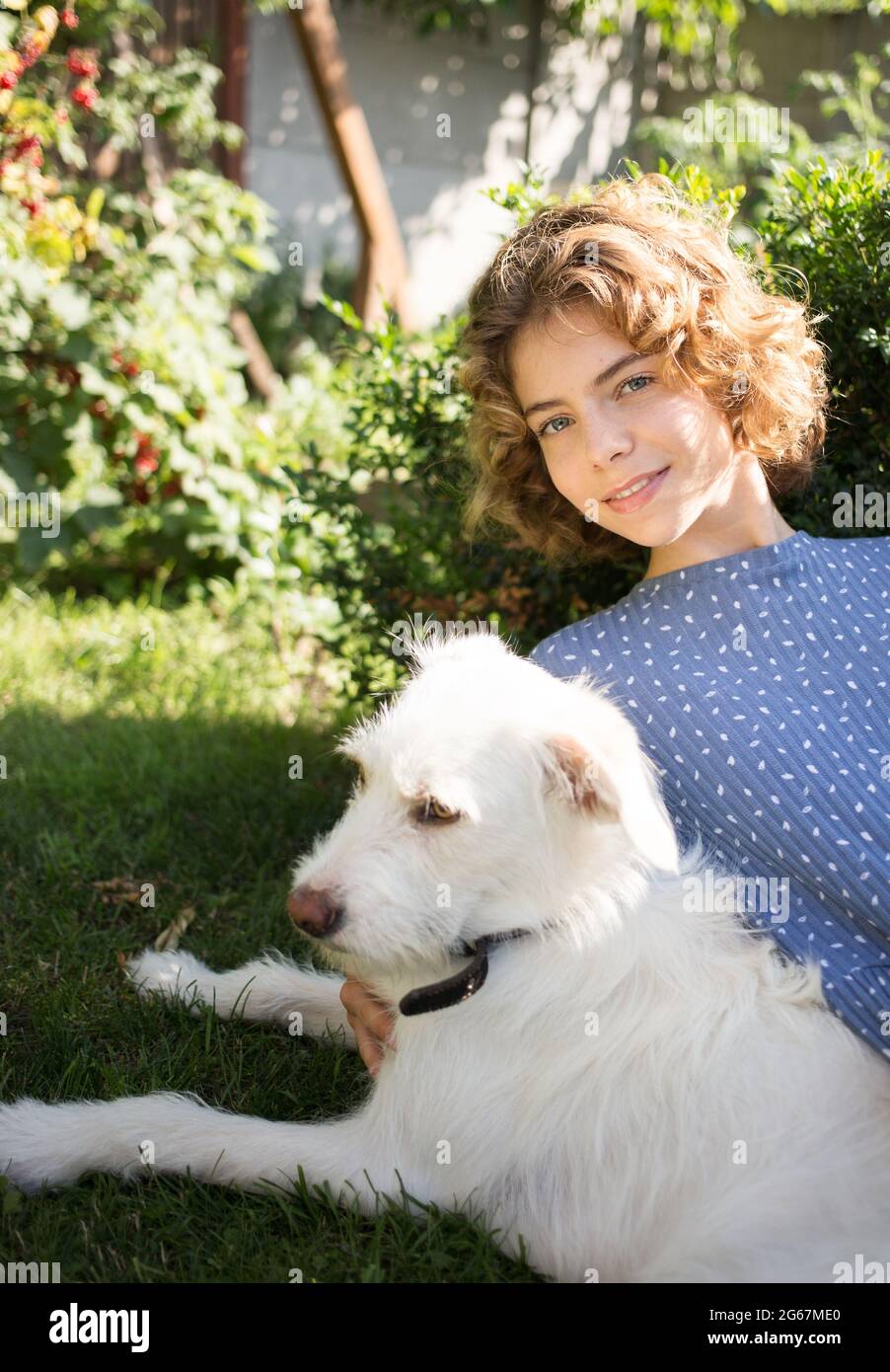 portrait of cute girl - teenager 17 - 18 years old lies on green grass next to her white dog. Focus on young woman. Summer relaxation with favorite pe Stock Photo