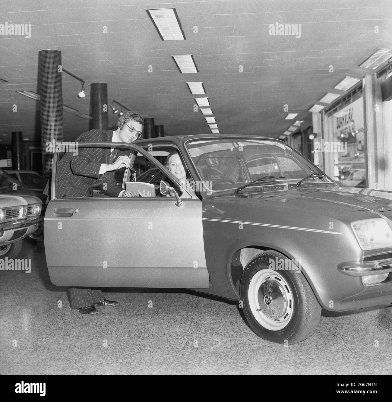 1975, historical, inside a car showroom, a salesman in a pin-stripe suit holding the keys and logbook of a new motorcar, a Vauxhall Chevette, with a lady customer -the new owner - sitting in the driver's seat, England, UK. A small family car, the Chevette was manufactured by Vauxhall motors in the UK between 1975 to 1984. With a design based on the Opel Kadett, the Chevette was one of the first British-built hatchbacks and from 1975 to 1978, it was the UK's best-selling model in that class. Stock Photo