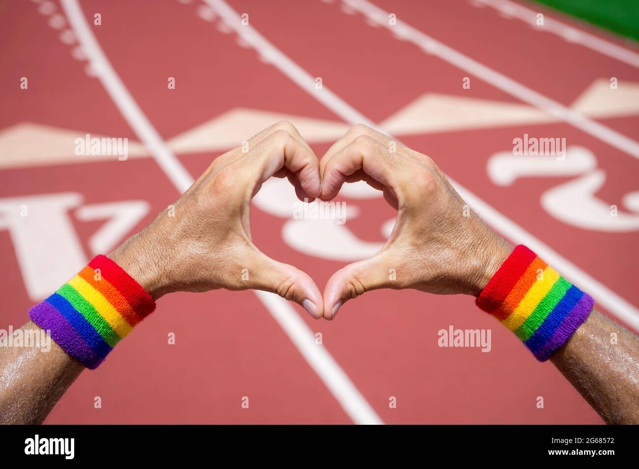 Gay athlete wearing rainbow pride wristbands making love heart hands gesture against a red athletic track background Stock Photo