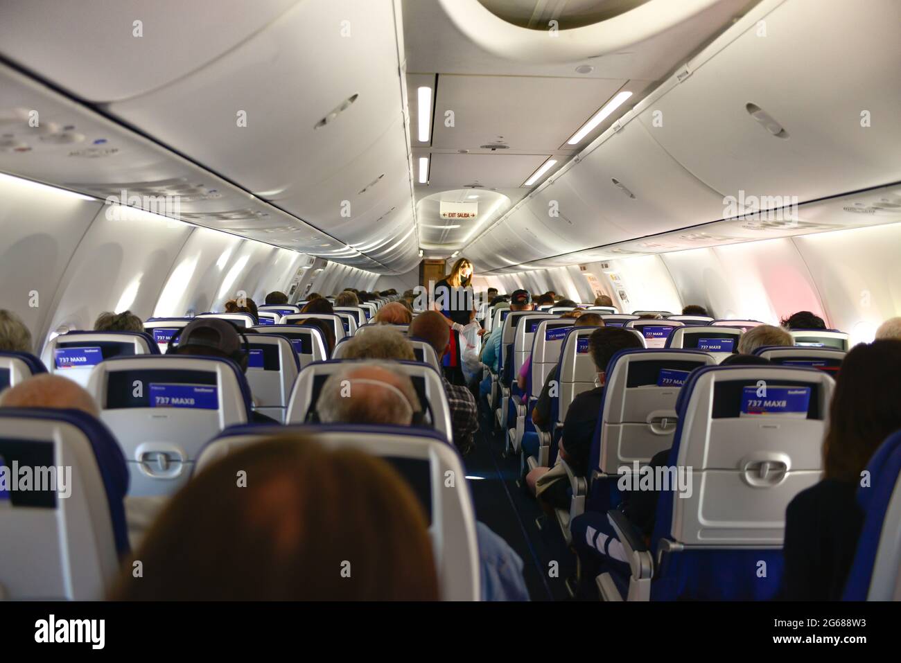 View from the Back of the plane of flight attendant in cabin of a commercial 737 Max airplane loaded with passengers, the Max newly returning to servi Stock Photo