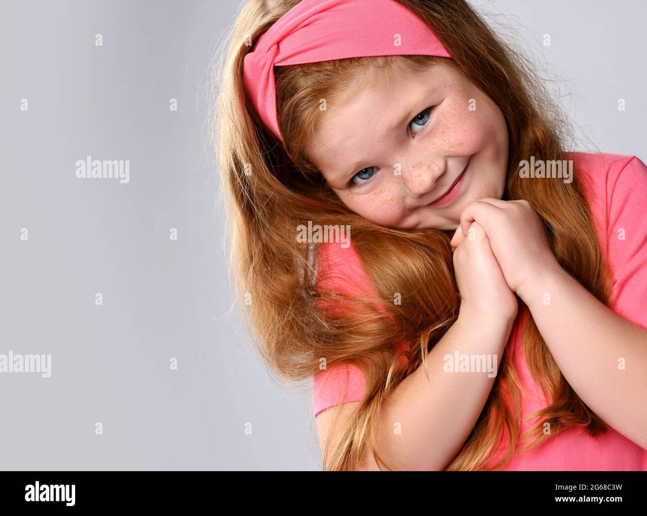 Portrait of cute, sly smiling redhead kid girl with freckles on face in pink t-shirt and headband holding hands together Stock Photo