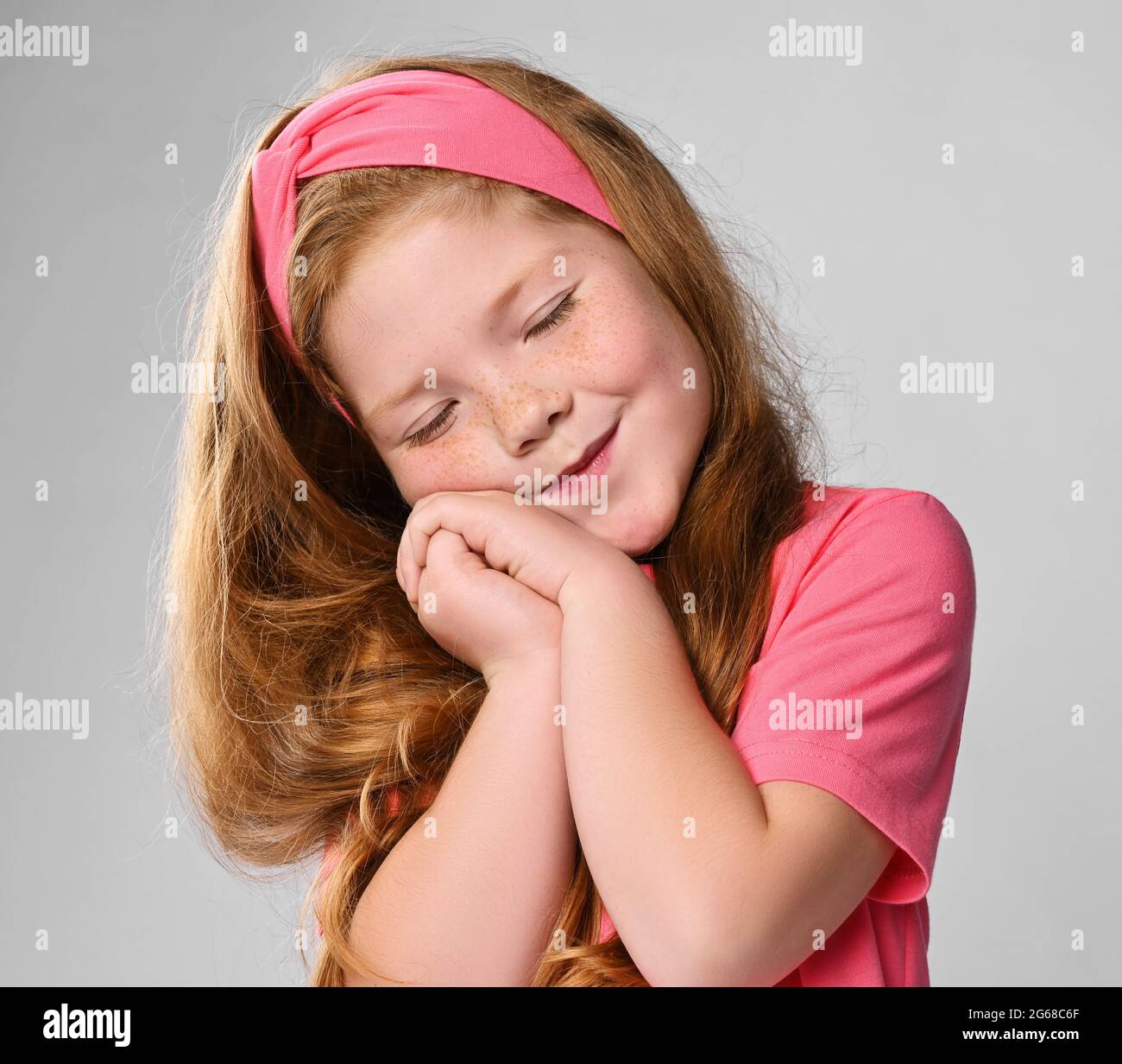 Portrait of cute daydreaming redhead kid girl in pink t-shirt and headband holding hands together, eyes closed Stock Photo